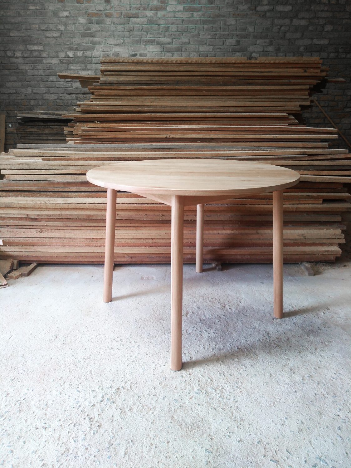 Pale wooden table with a round top and round section legs with a stack of timber in the background.