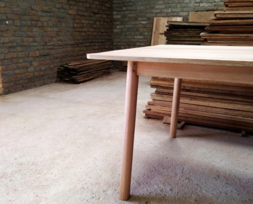 Pale wooden table leg detail with round section legs, stacked timber in the background.