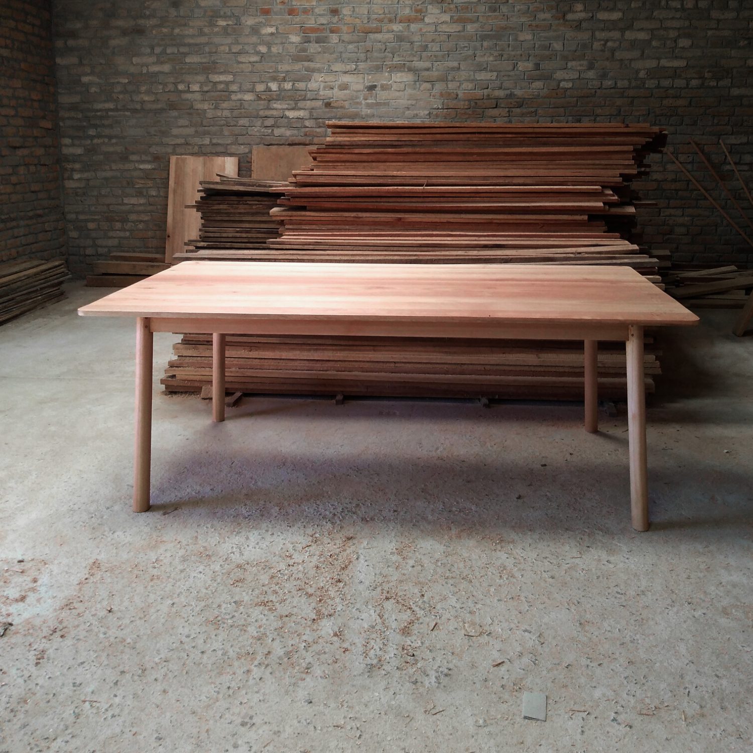 Pale wooden table with round section legs, stacked timber in the background.