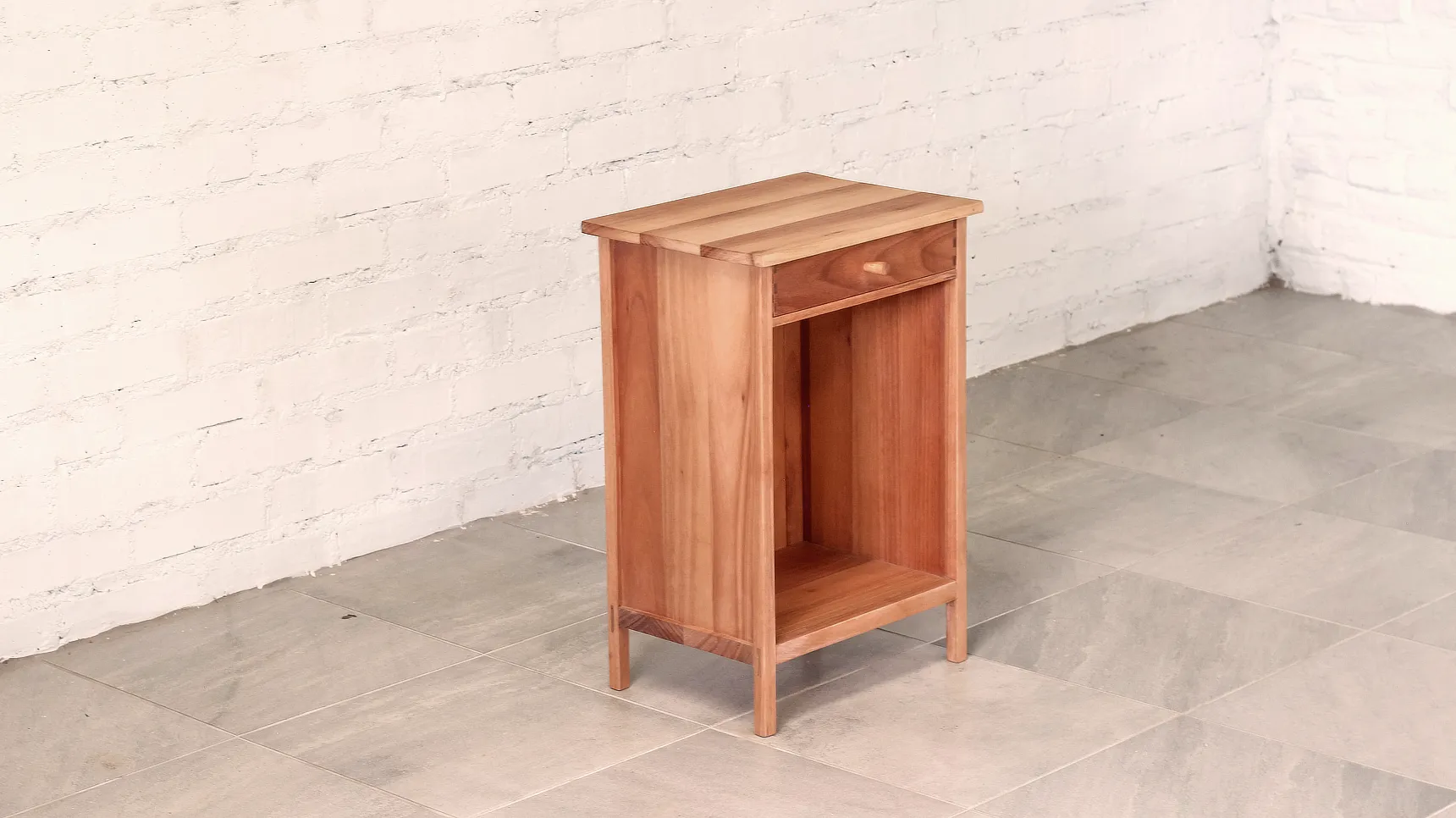 Bedside table made from pale brown timber with top drawer and open boxed construction with legs