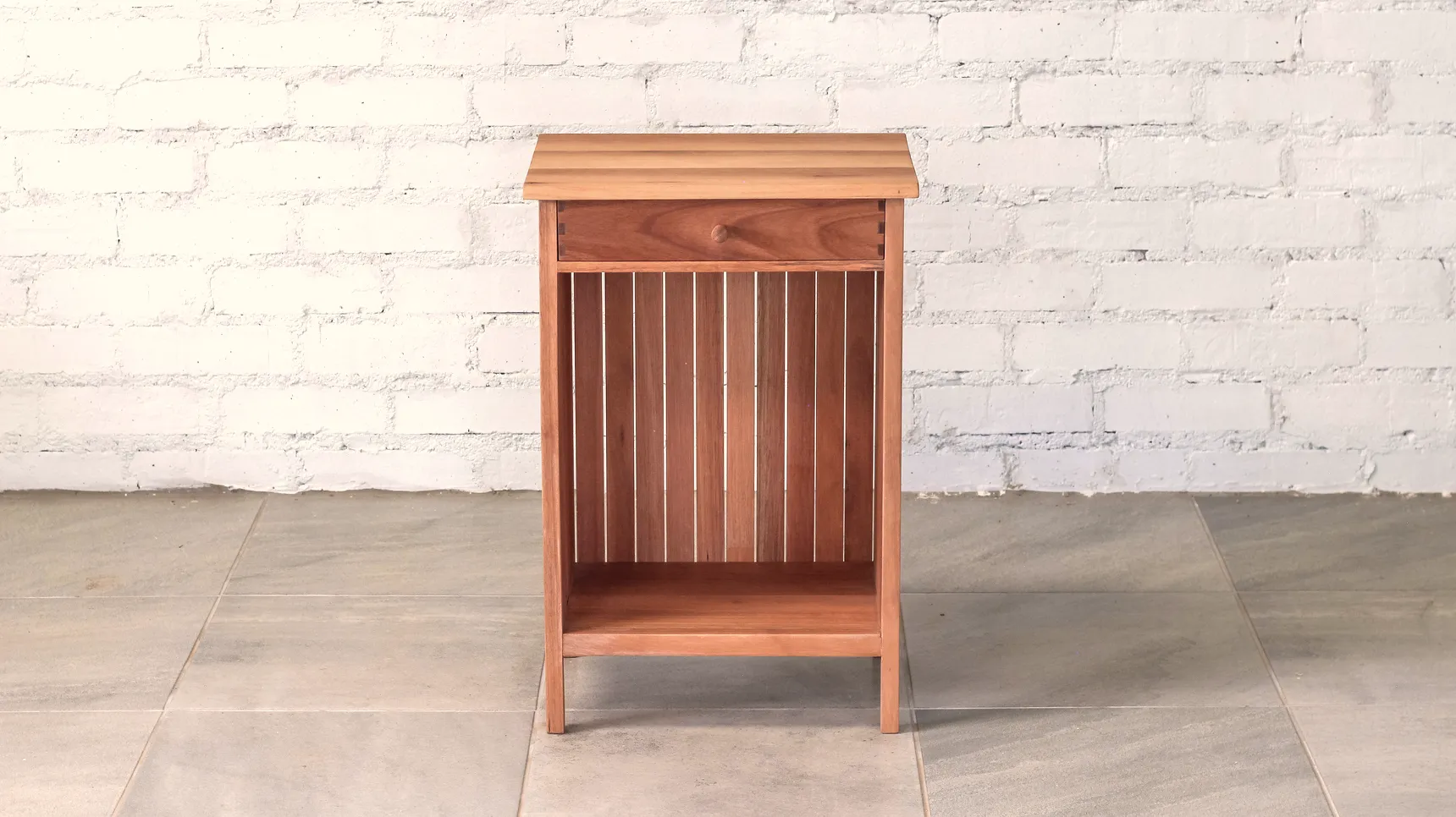 Bedside table made from pale brown timber with top drawer, open boxed storage space and slatted hardwood back panel.