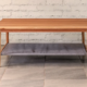 Coffee table made from a pale brown timber with round section gently splayed legs and a fabric shelf.